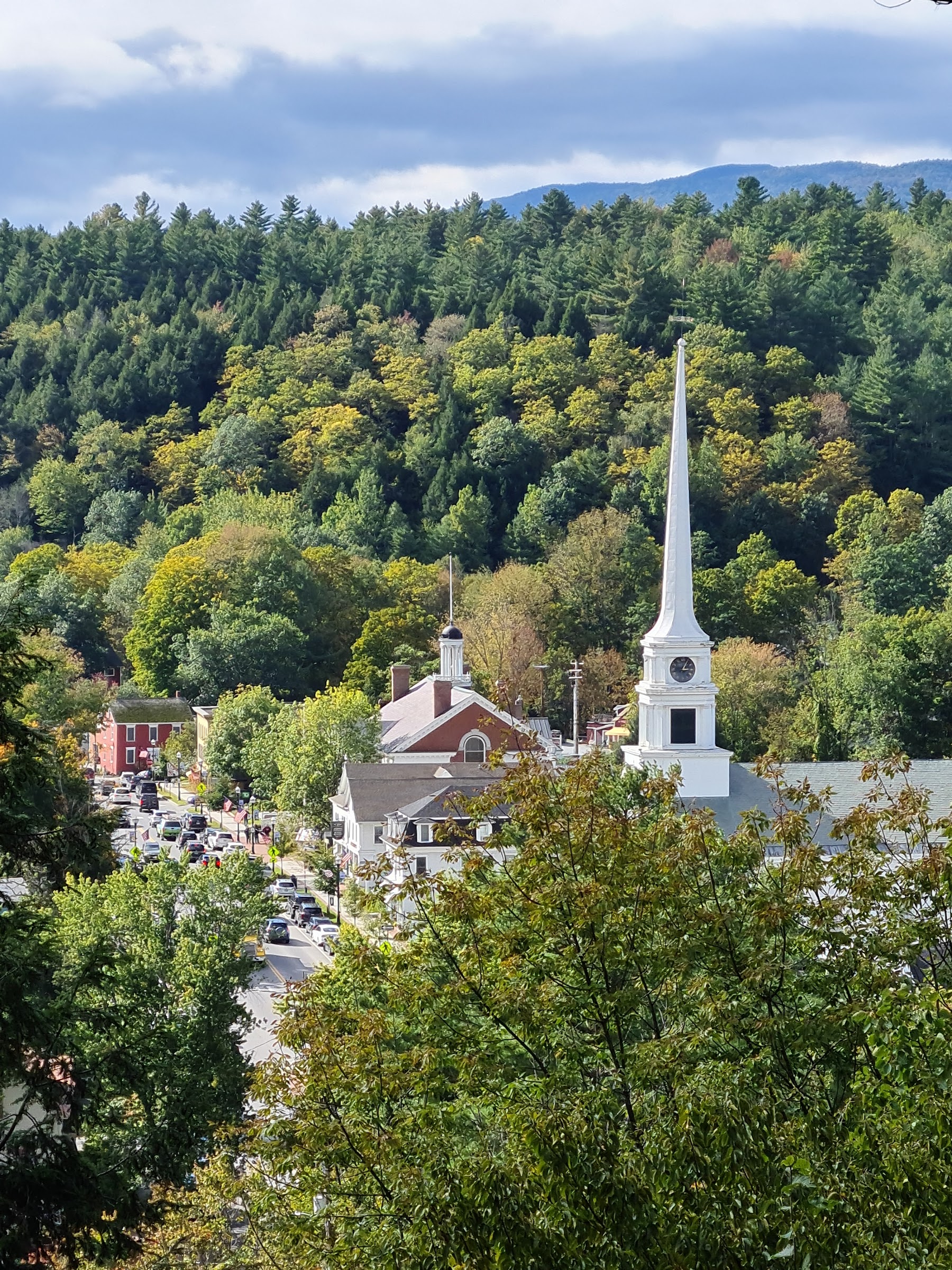 A quaint white church with a tall steeple stands against a backdrop of vibrant green Vermont hills.
